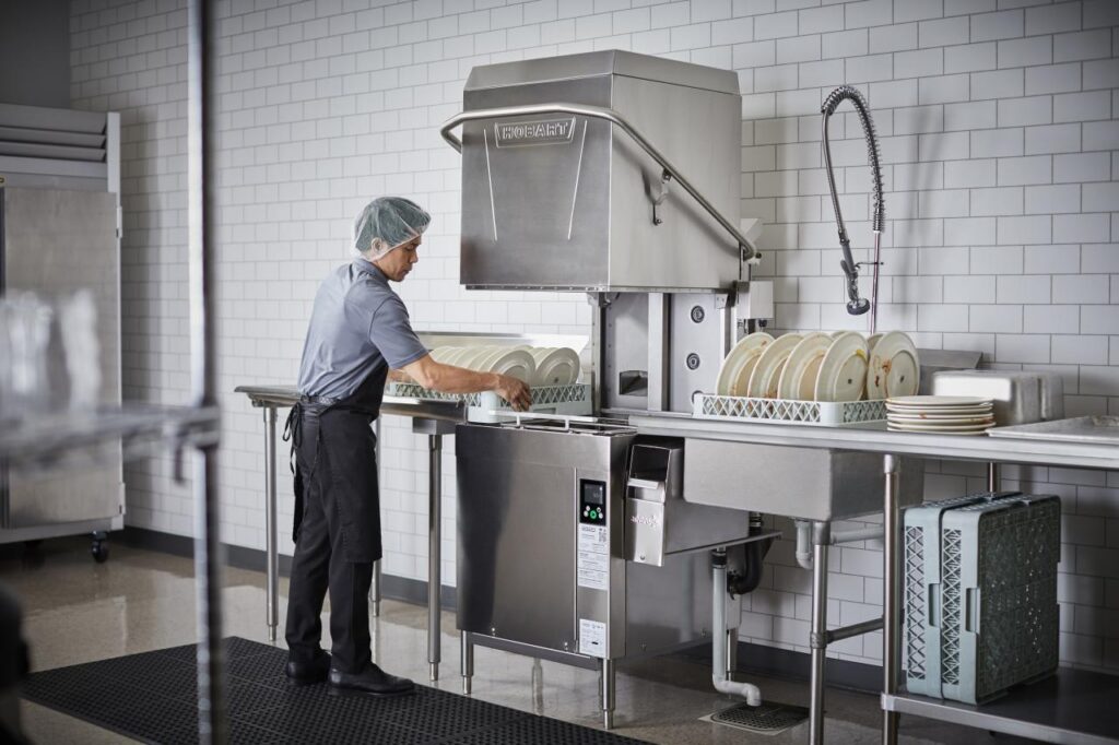 Worker operating a commercial dishwasher, illustrating the equipment covered by dish chemical and service vendor programs.