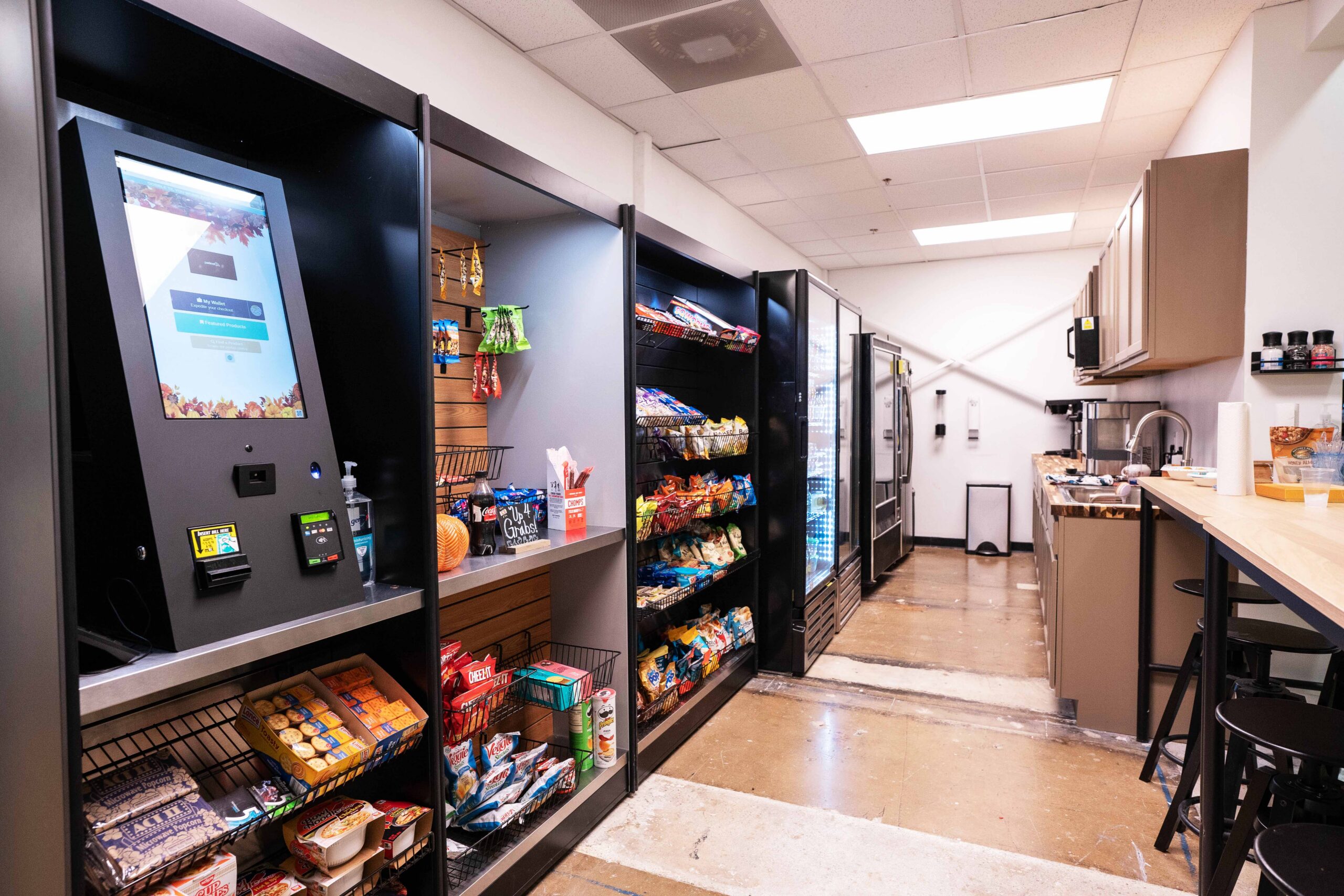 Self-checkout kiosk and open shelving at an office micro market, showing the self-service payment terminal and product range Denver Beverage manages remotely