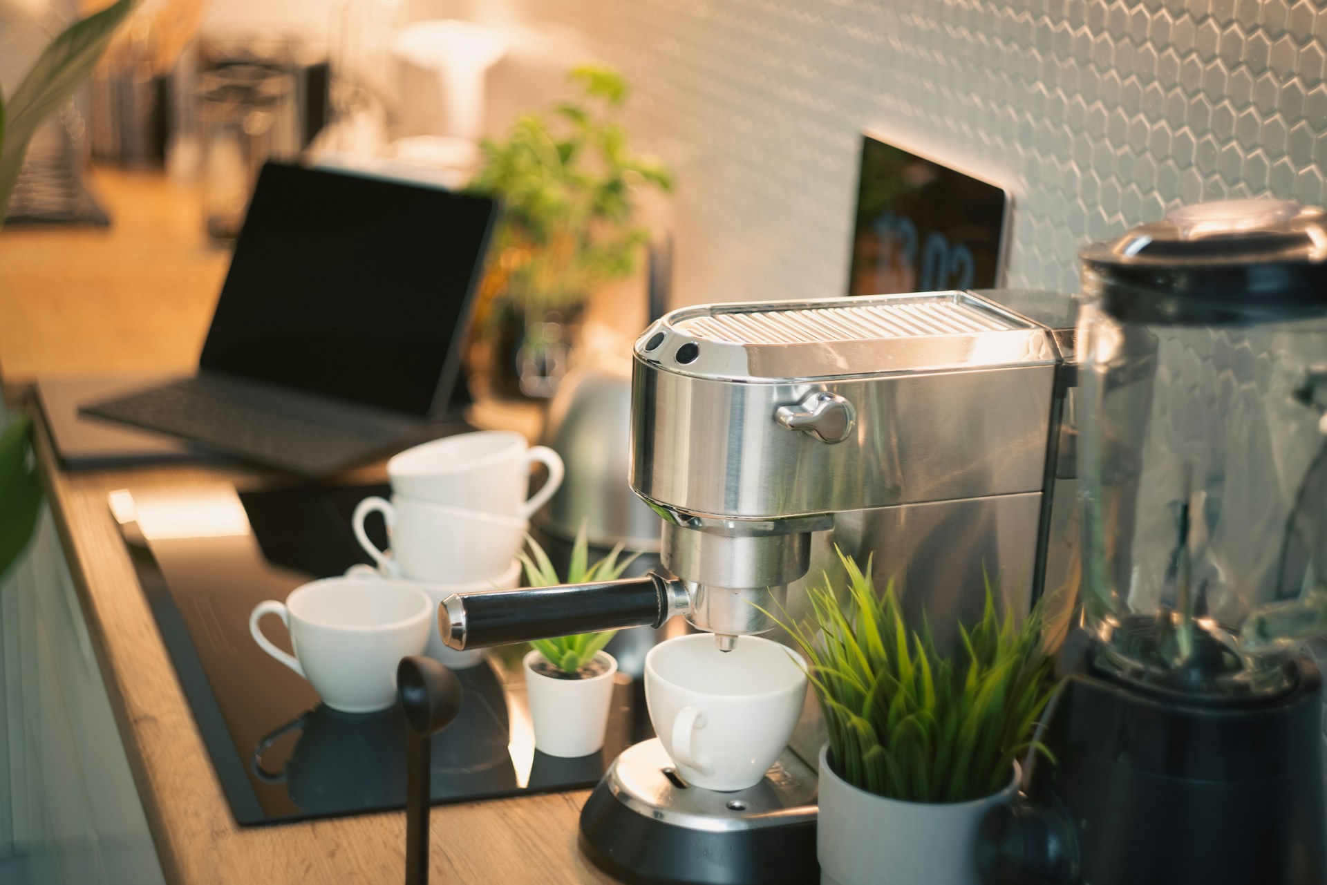 Compact stainless espresso machine with white cups on an office desk beside a laptop and plants