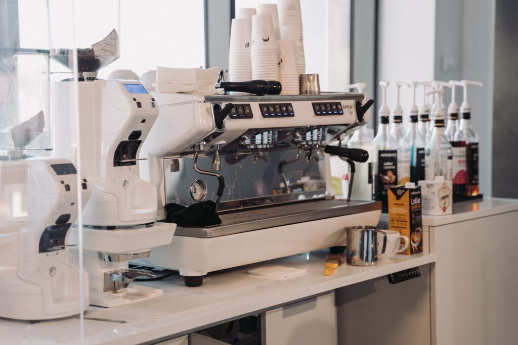 White commercial espresso machine and grinder installed on a bright office counter with syrup bottles and cups
