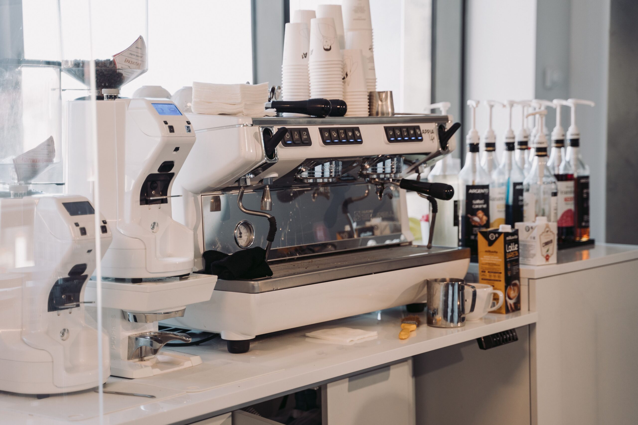 White commercial espresso machine and grinder installed on a bright office counter with syrup bottles and cups