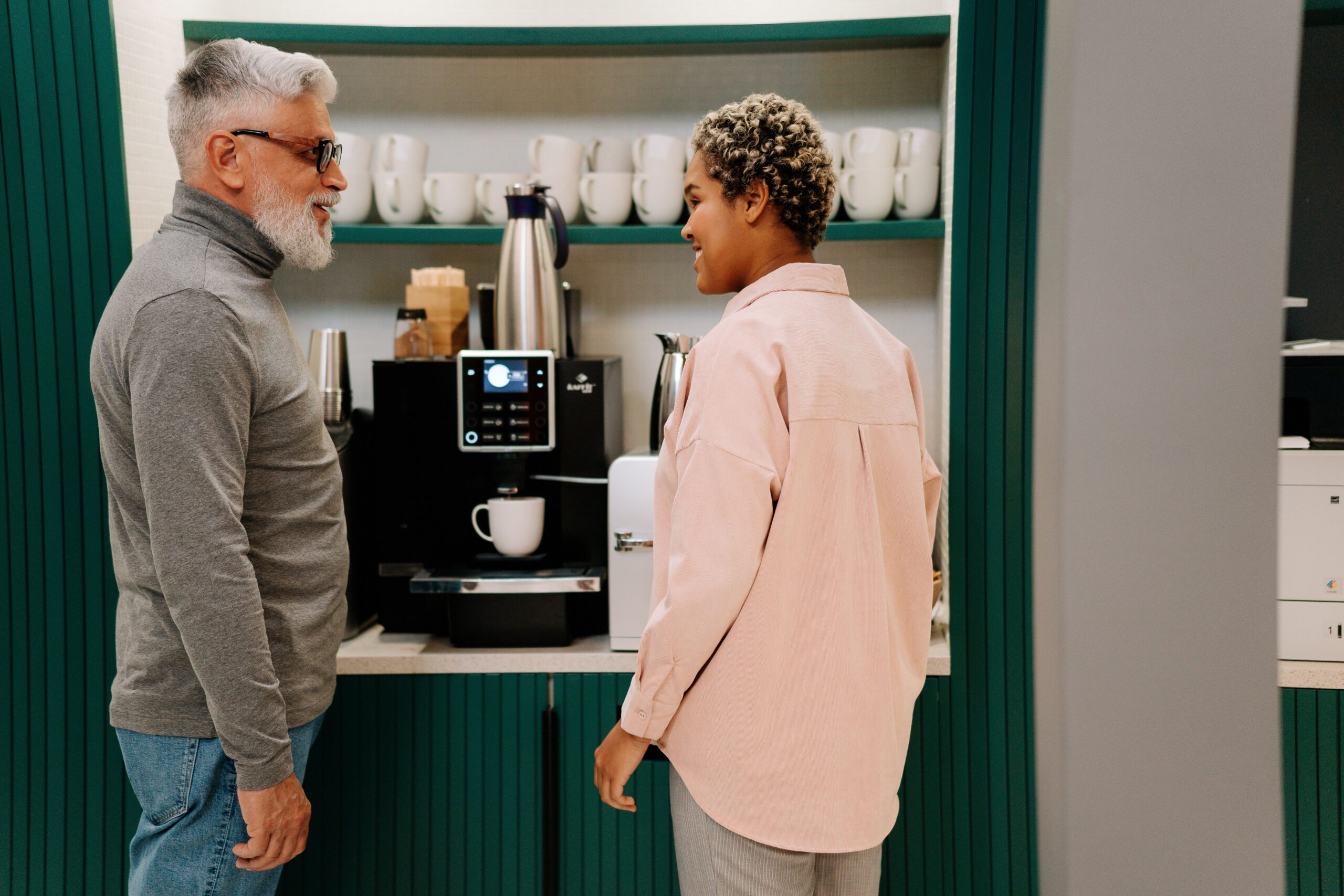 Older man and staff member chat beside a bean-to-cup coffee station in a senior living community lounge