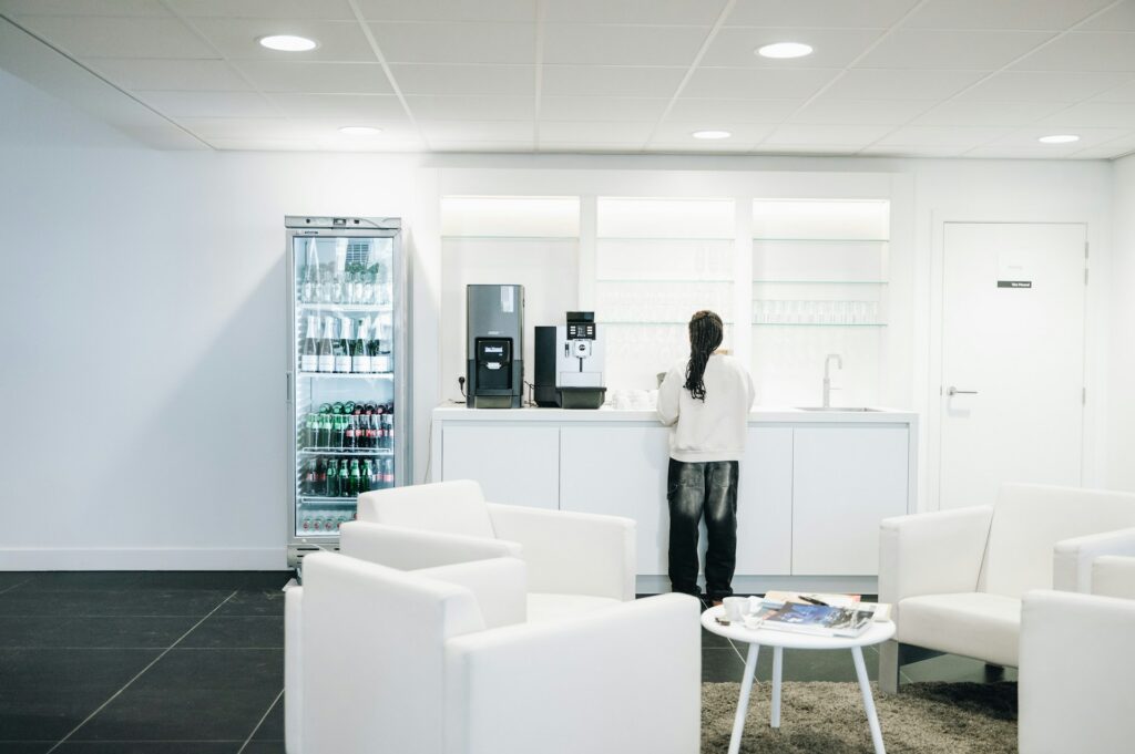 Person at a coffee station beside a stocked beverage cooler in a hospital waiting area with white armchairs.