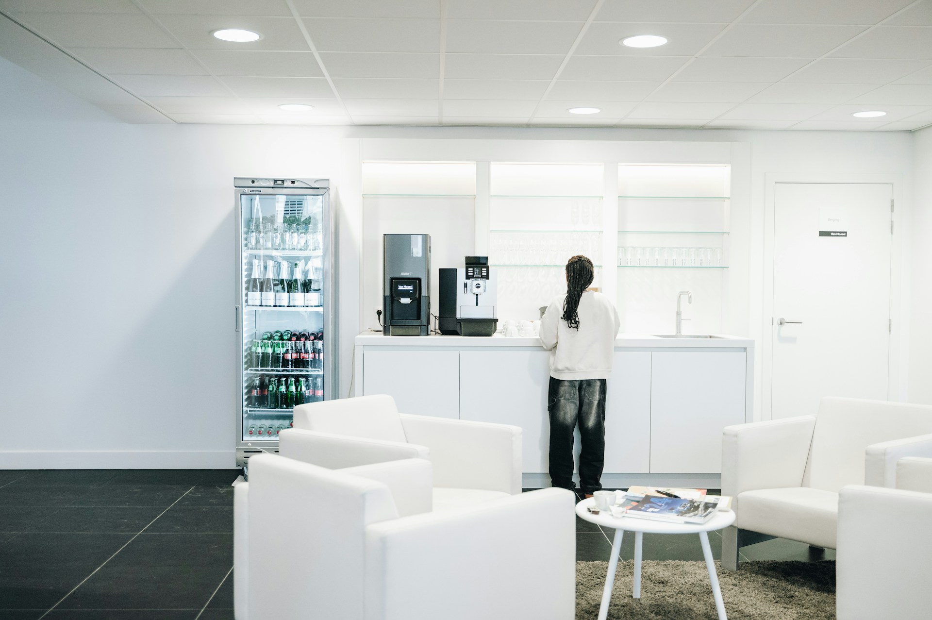 Person at a coffee station beside a stocked beverage cooler in a hospital waiting area with white armchairs.