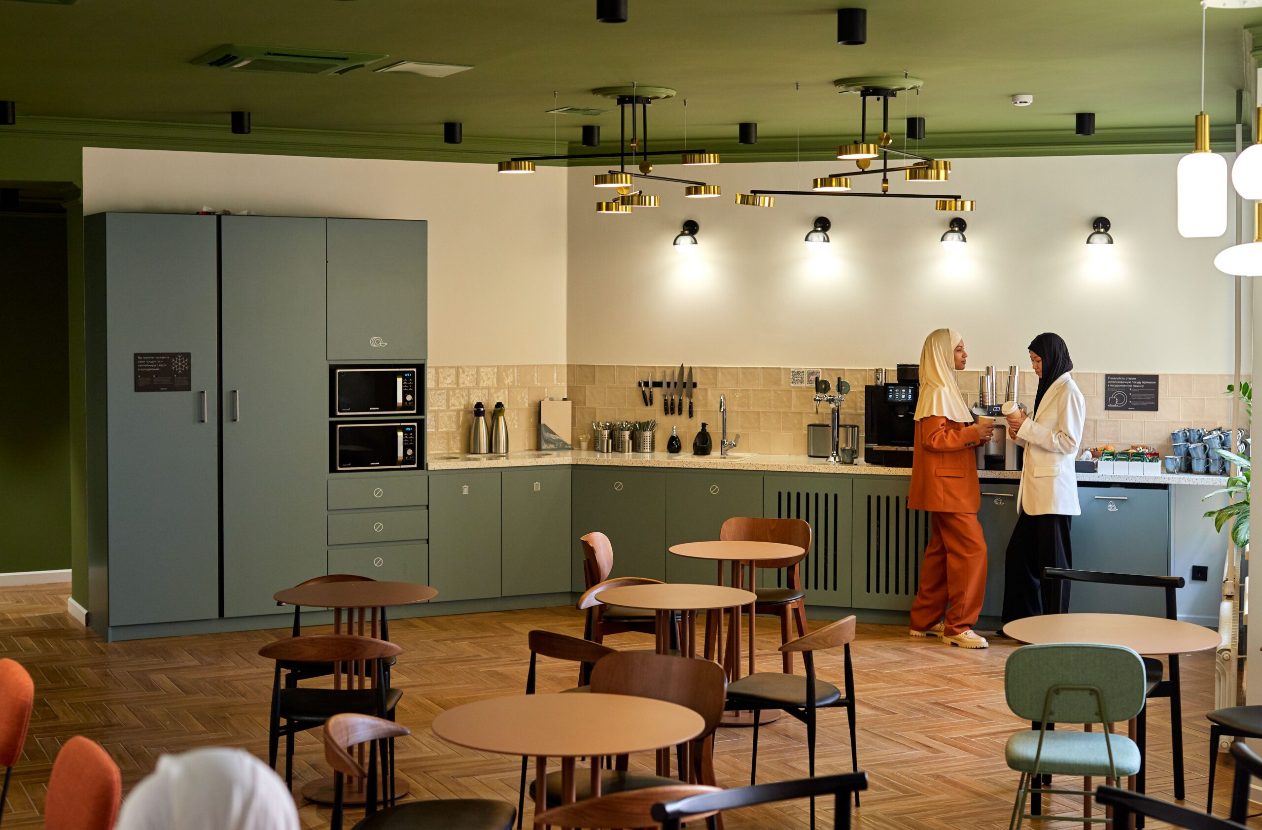 Two coworkers chat by the coffee station in a modern green office breakroom with tables and chairs