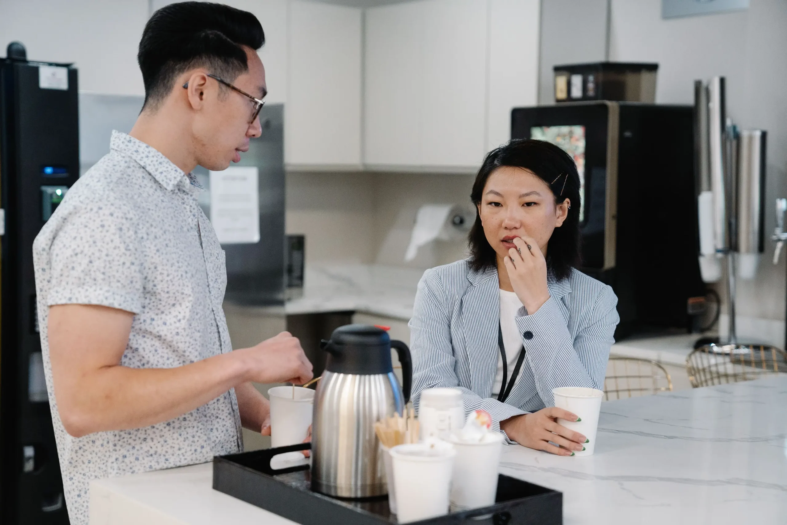 Two coworkers chatting over cups in an office breakroom with a commercial coffee machine and thermal carafe.