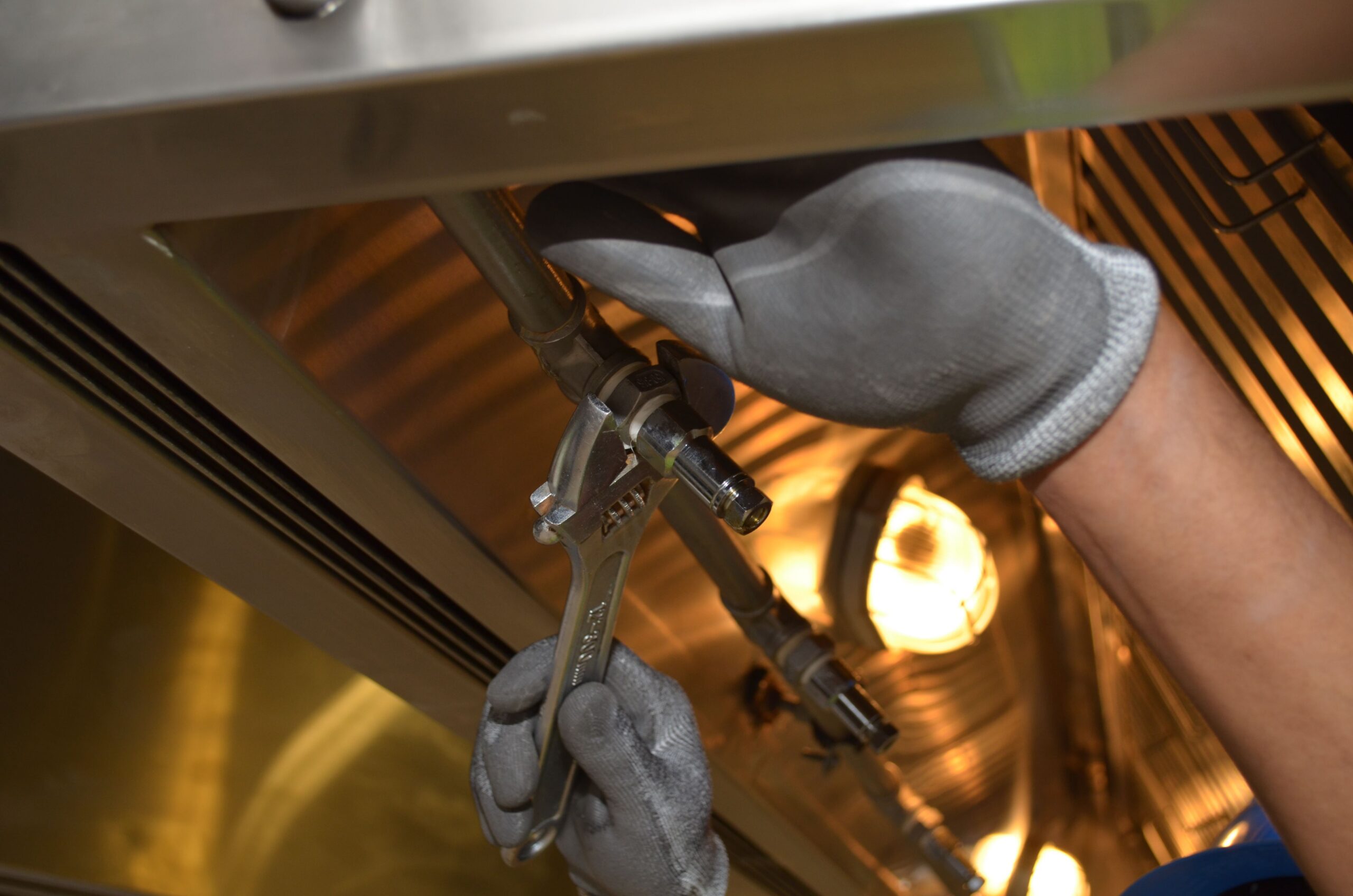 Gloved technician tightening a gas line fitting under stainless commercial kitchen equipment during scheduled maintenance