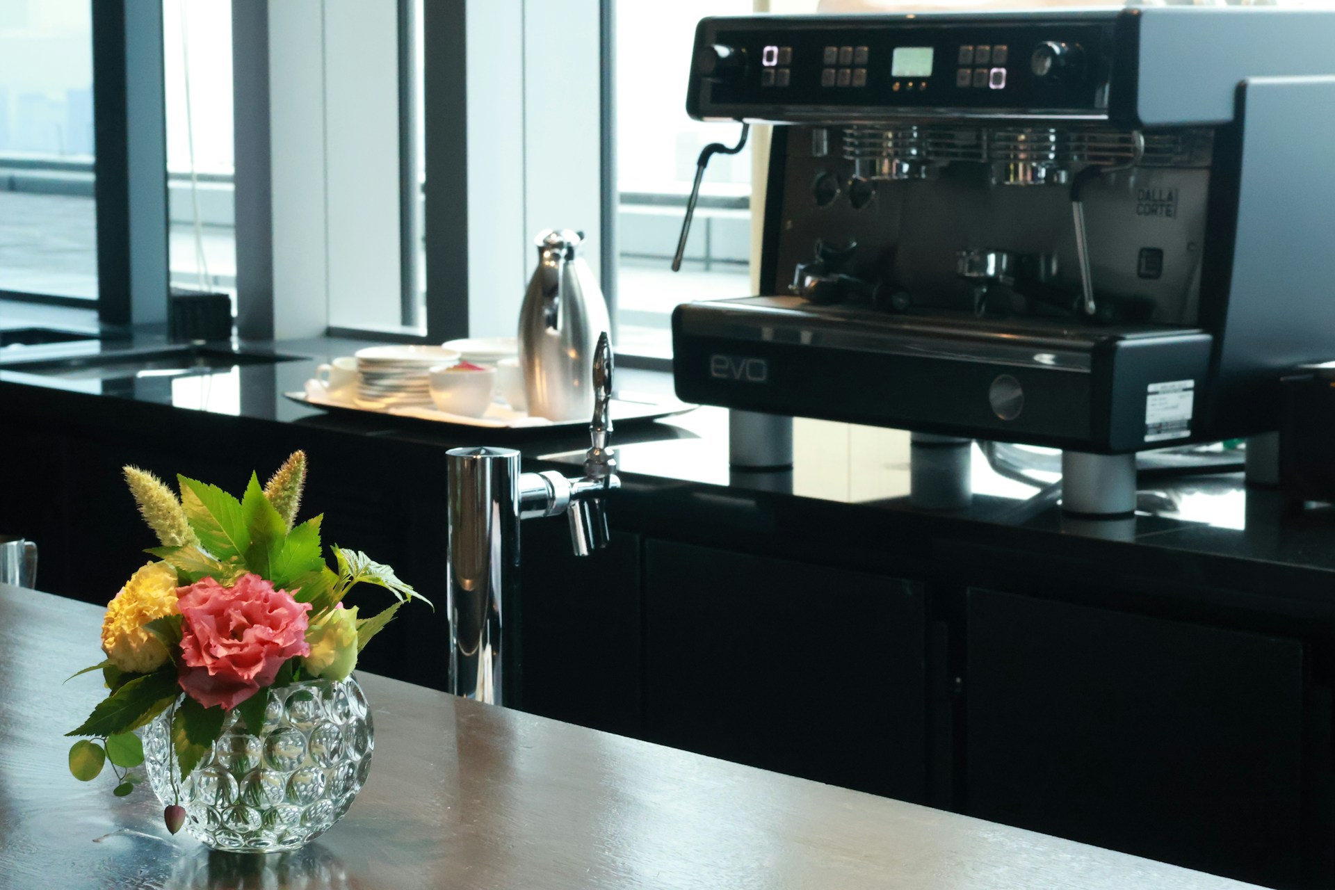 Unbranded commercial espresso machine on a hotel lobby counter beside a floral arrangement and window wall